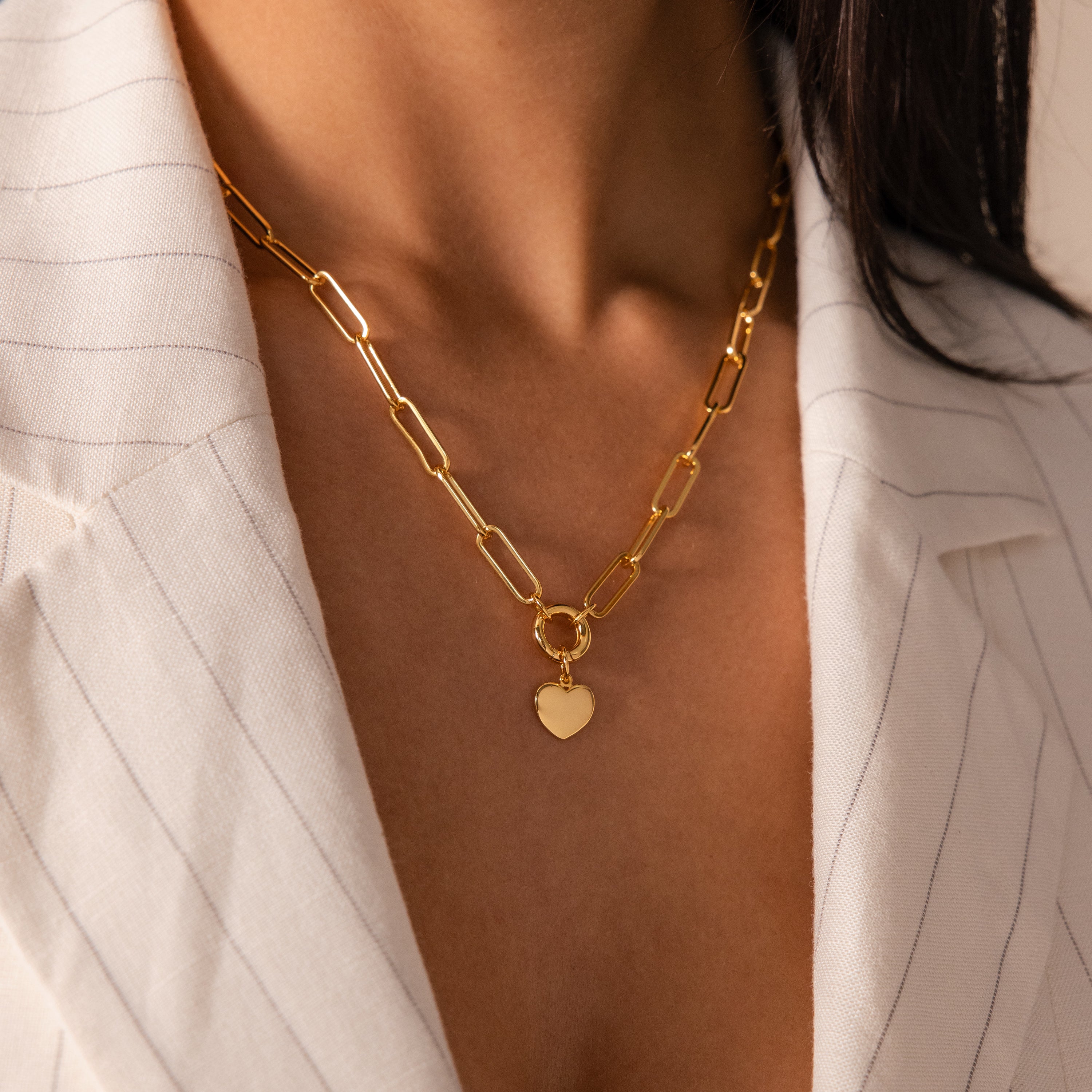 A woman wearing the Paperclip Heart Charm Necklace in 18K Gold with a heart pendant over a white pinstripe blazer.