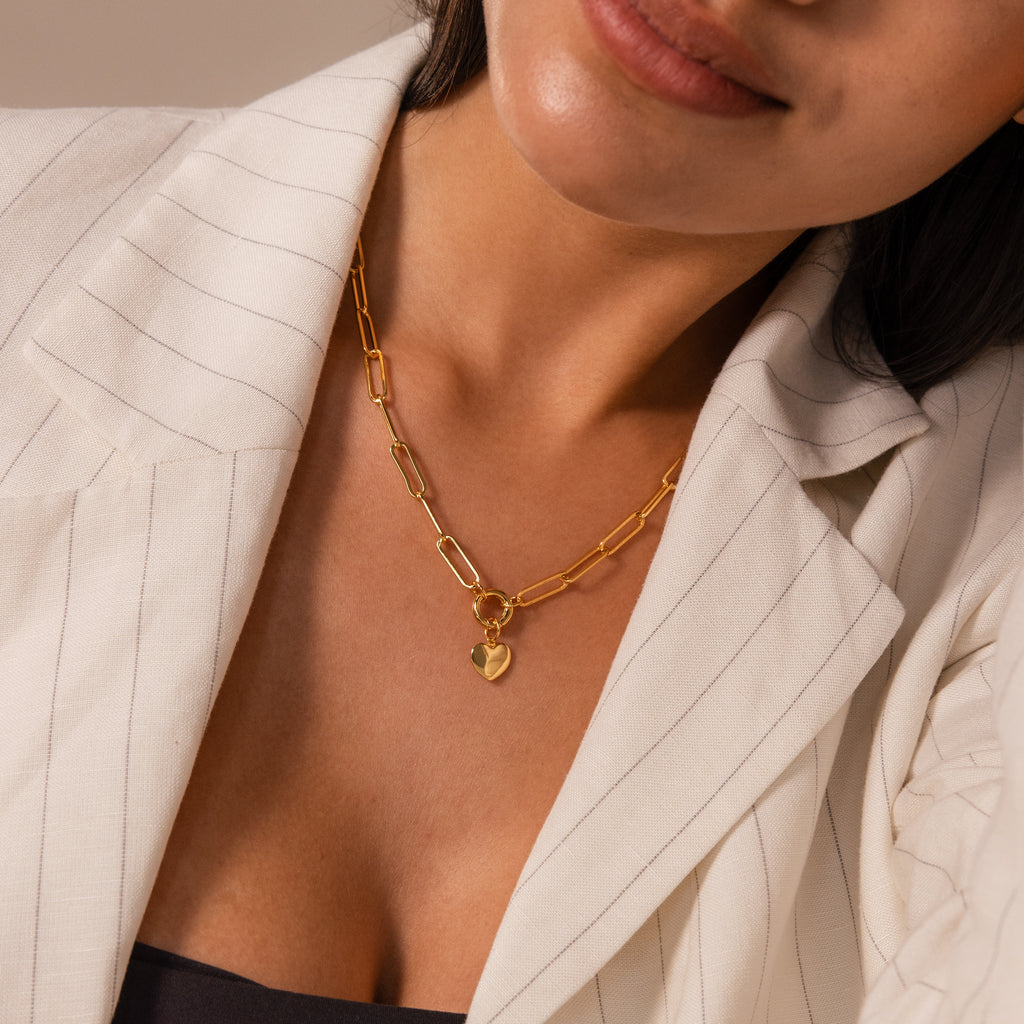 A woman in a white pinstripe blazer wears the Paperclip Heart Charm Necklace in 18K Gold, featuring a heart pendant.
