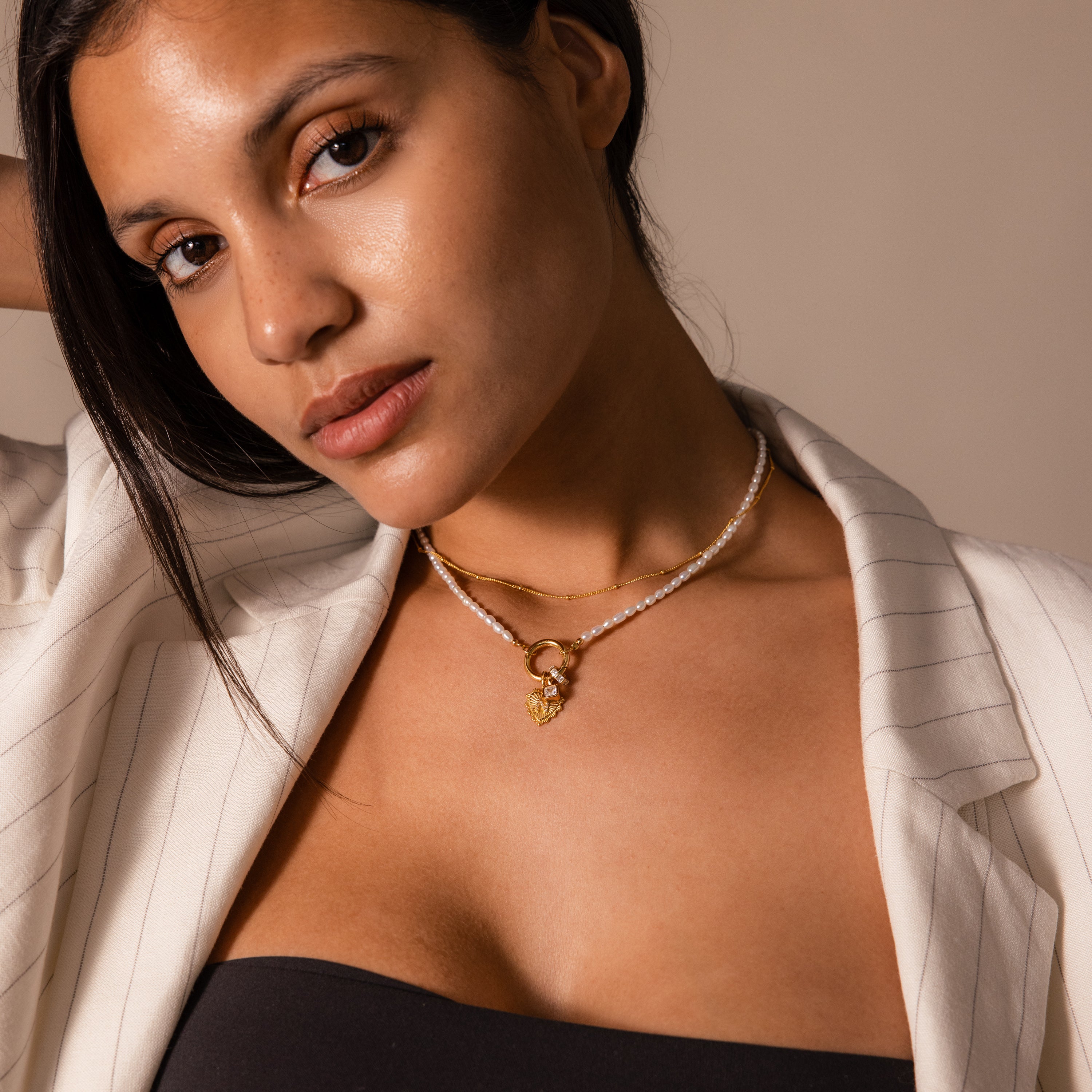 A woman in a pinstriped blazer and black top wears layered gold necklaces, featuring the Pearl Chain Clasp Necklace, while posing with her hand behind her head.