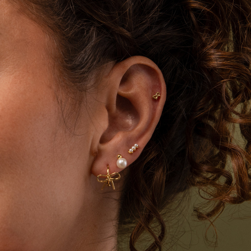 Close up of a model wearing an earring stack with our Tiny Bow Flatback Studs in Gold, Bow Huggies and Pearl Studs