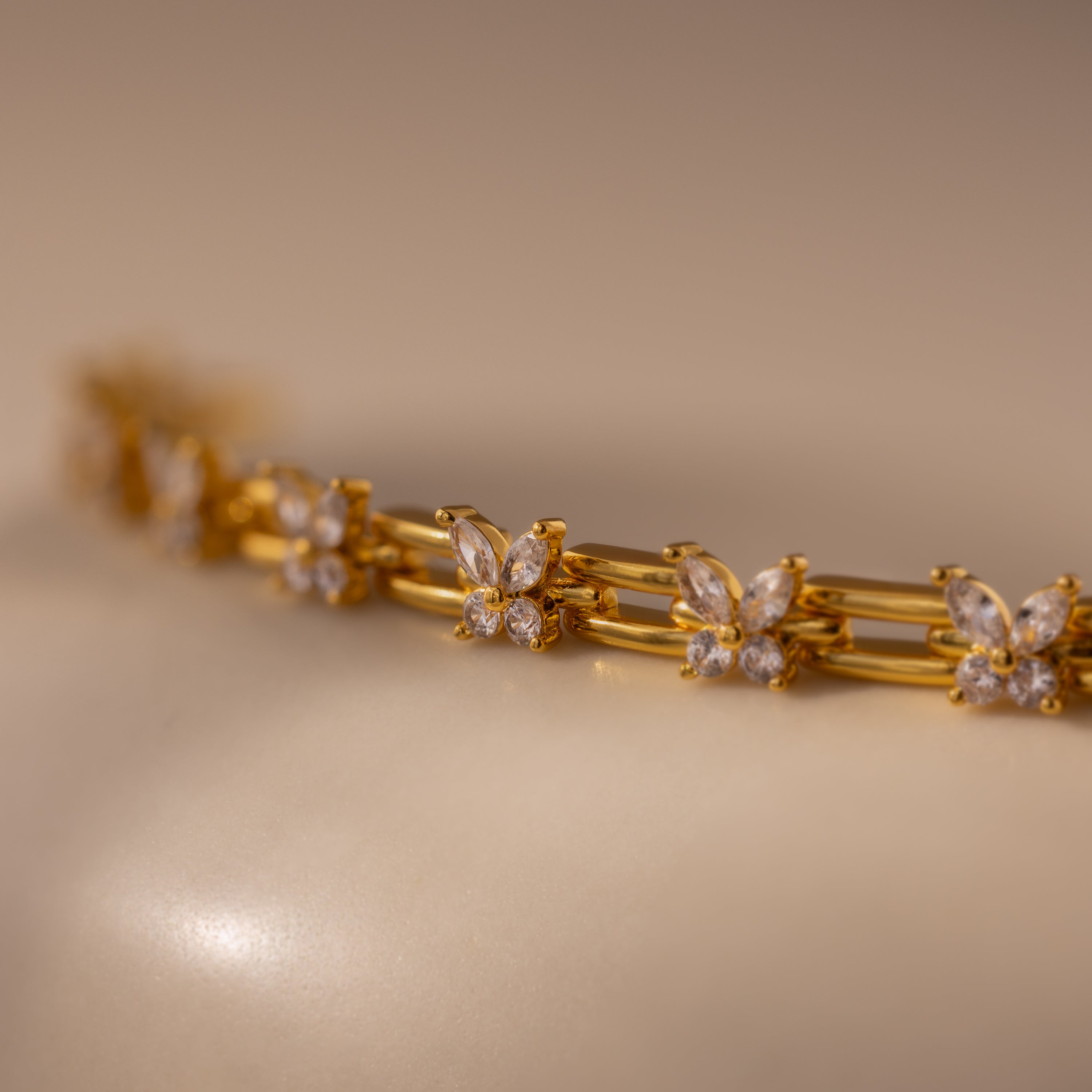 The Mariposa Link Bracelet, a gold piece adorned with diamond settings, rests elegantly on a smooth beige surface—a stunning choice for feminine jewelry.