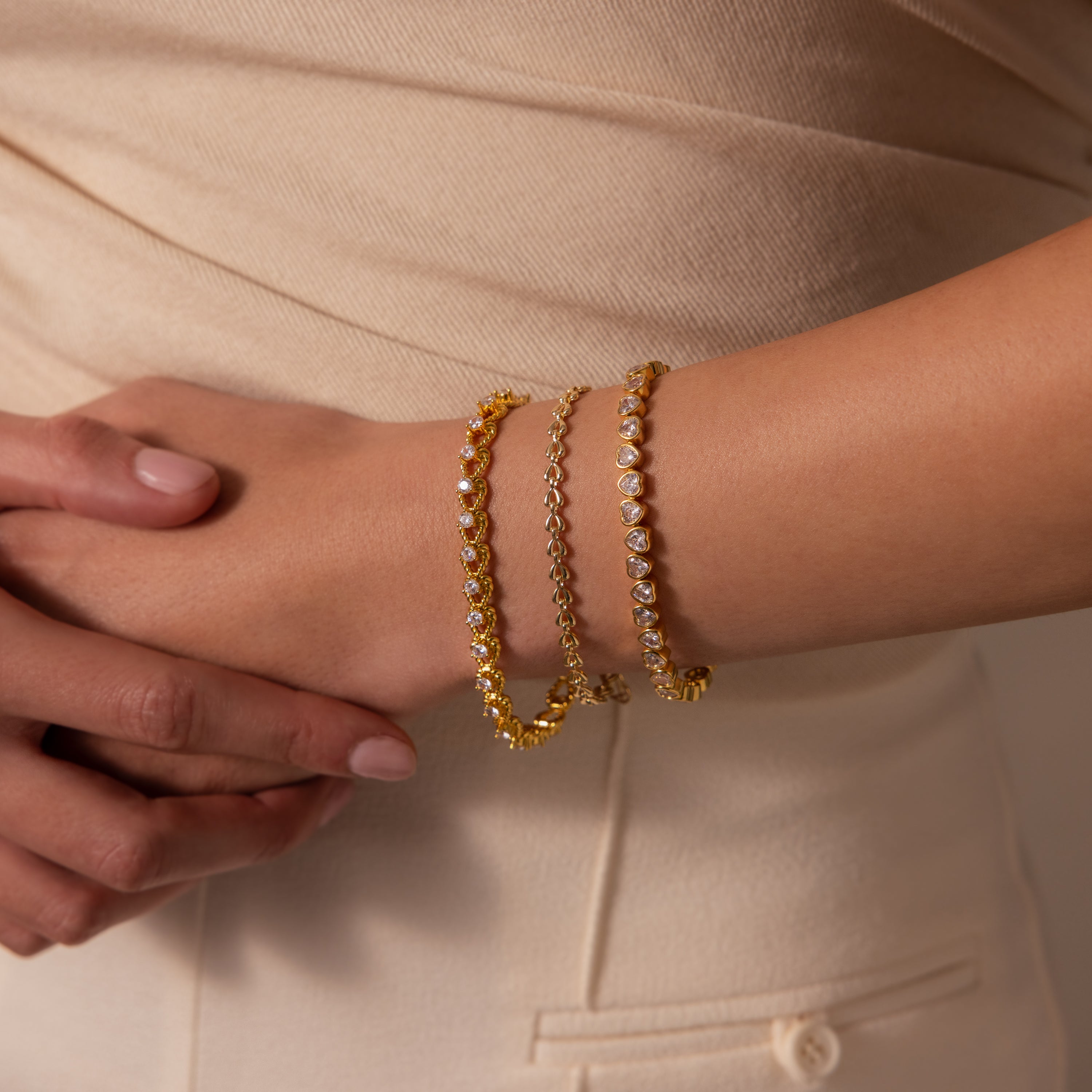 A woman in beige clothing wears elegant jewelry, showcasing the Heart Tennis Bracelet in 18K Gold as she rests her hand on her hip.