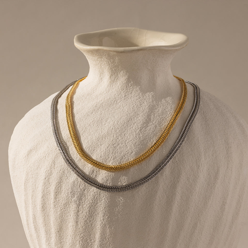 A Thin Mesh Chain Necklace in 18K Gold is draped on a white ceramic vase against a neutral background.
