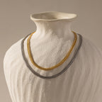 A Thin Mesh Chain Necklace in 18K Gold is draped on a white ceramic vase against a neutral background.