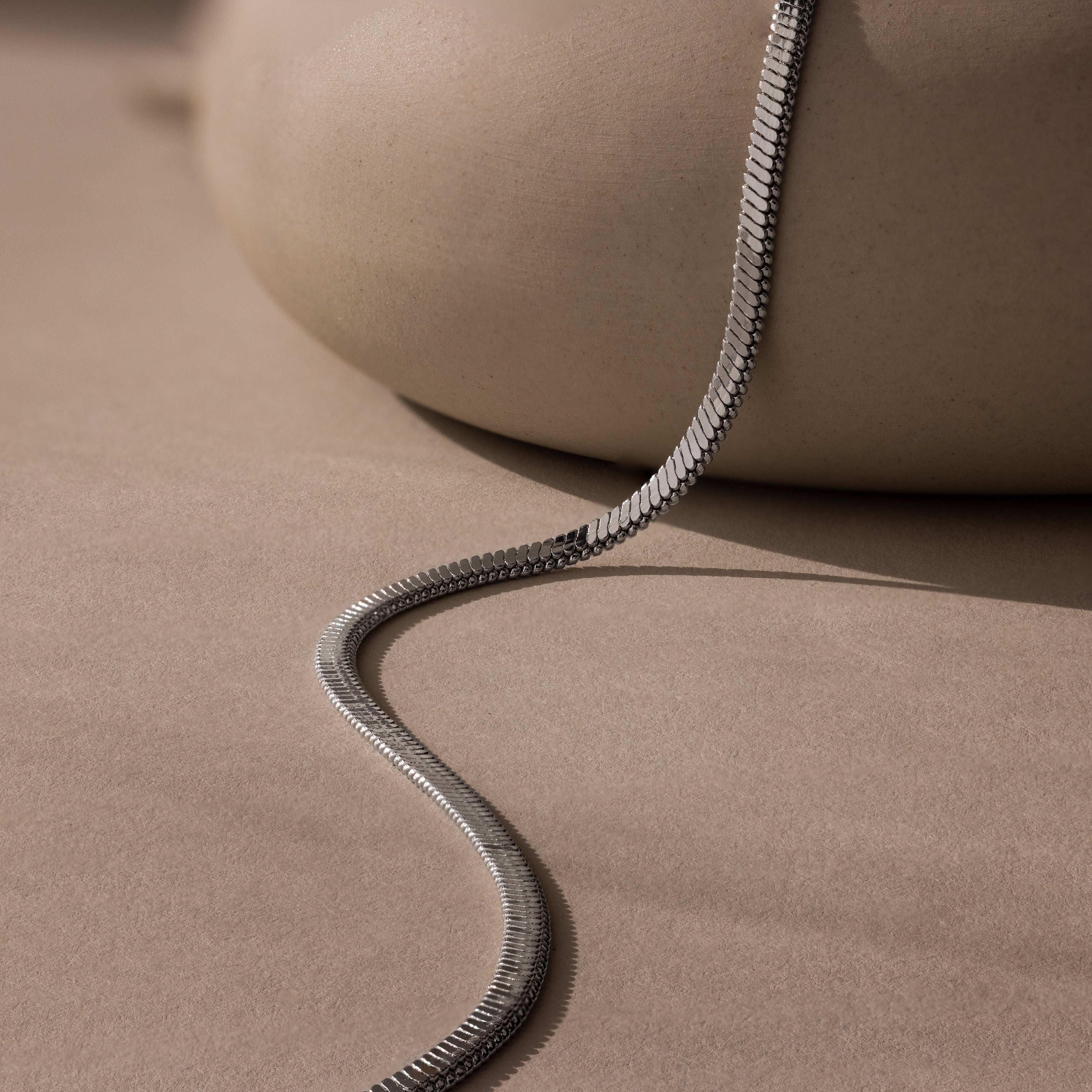 A modern jewelry piece, the Bold Snake Chain Necklace rests on a smooth beige surface with soft shadows—perfect as a sleek layering necklace.
