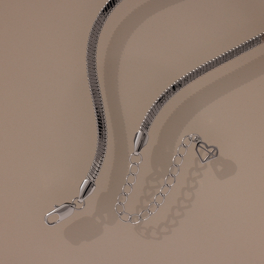 Modern jewelry at its finest—the Twisted Snake Chain Necklace in Silver features a smooth silver chain, clasp, and extender with an open heart charm, showcased on a beige background.