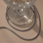 The Twisted Snake Chain Necklace in Silver is elegantly draped around the base of a clear glass on a beige surface, showcasing the refined beauty of modern jewelry.