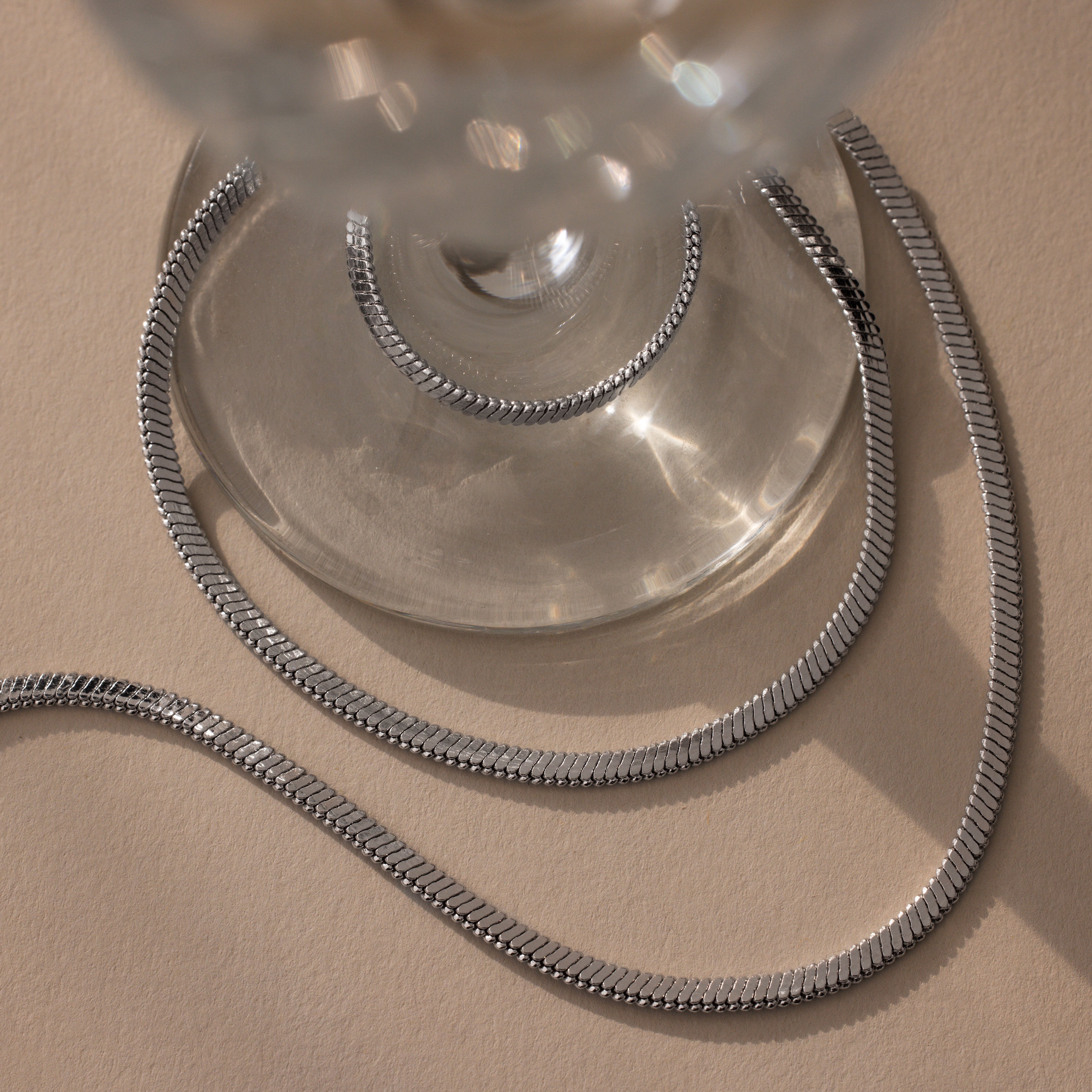 The Twisted Snake Chain Necklace in Silver is elegantly draped around the base of a clear glass on a beige surface, showcasing the refined beauty of modern jewelry.