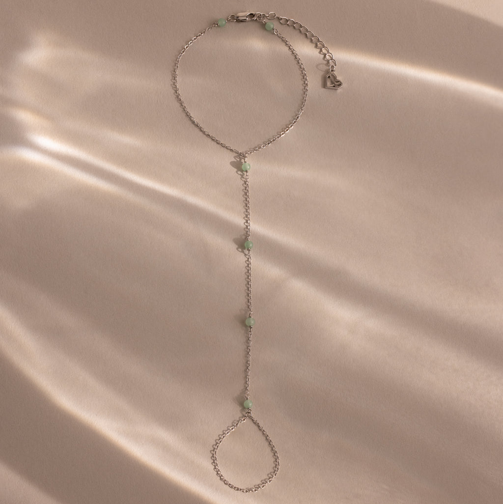 Jade Hand Chain Bracelet in Silver featuring small pale green jade beads, shown on a beige surface with soft, dappled light.
