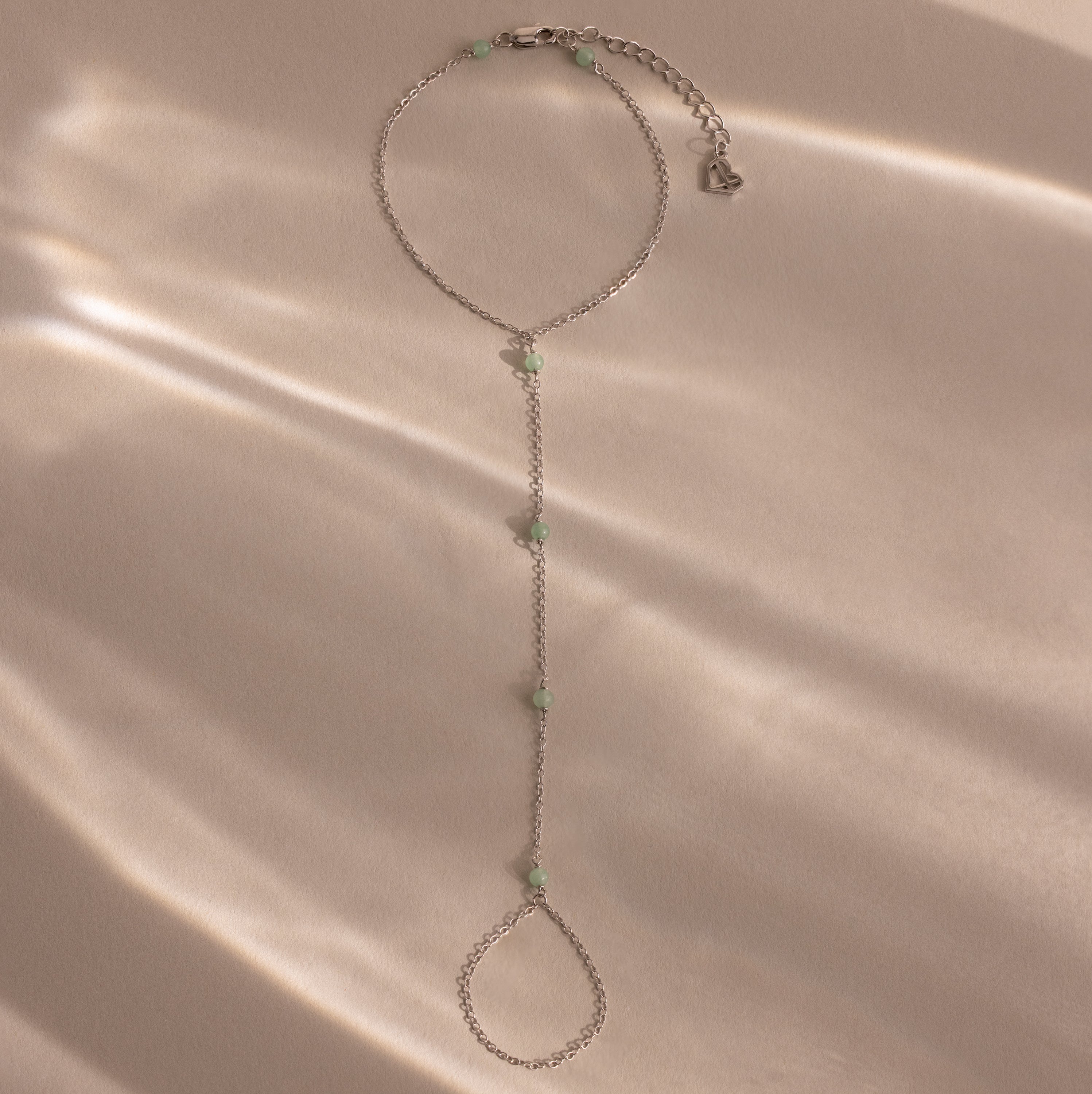 Jade Hand Chain Bracelet in Silver featuring small pale green jade beads, shown on a beige surface with soft, dappled light.