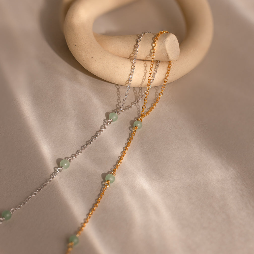 The Jade Hand Chain Bracelet features two delicate chains, one in silver and one in gold, each adorned with green jade beads and elegantly draped over a smooth cream-colored ring for a refined look.