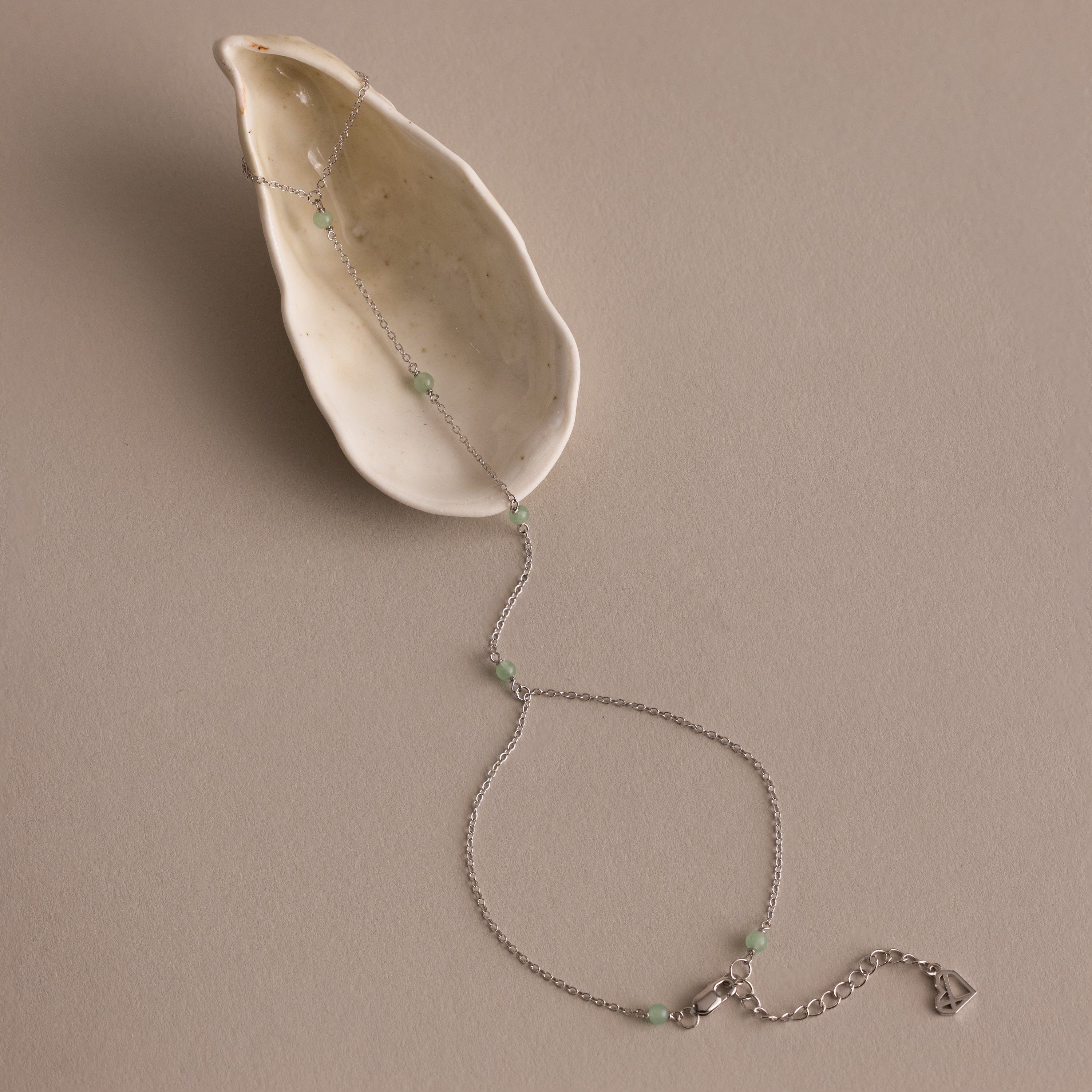 The Jade Hand Chain Bracelet in Silver is displayed on a cream-colored seashell, featuring a delicate silver chain adorned with jade beads.