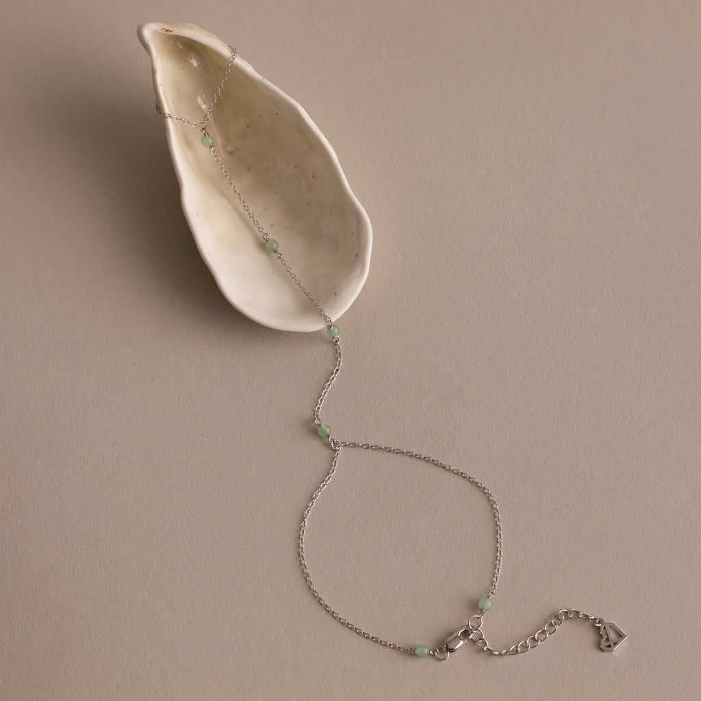 The Jade Hand Chain Bracelet features delicate silver and green jade beads, beautifully presented on a cream-colored shell.