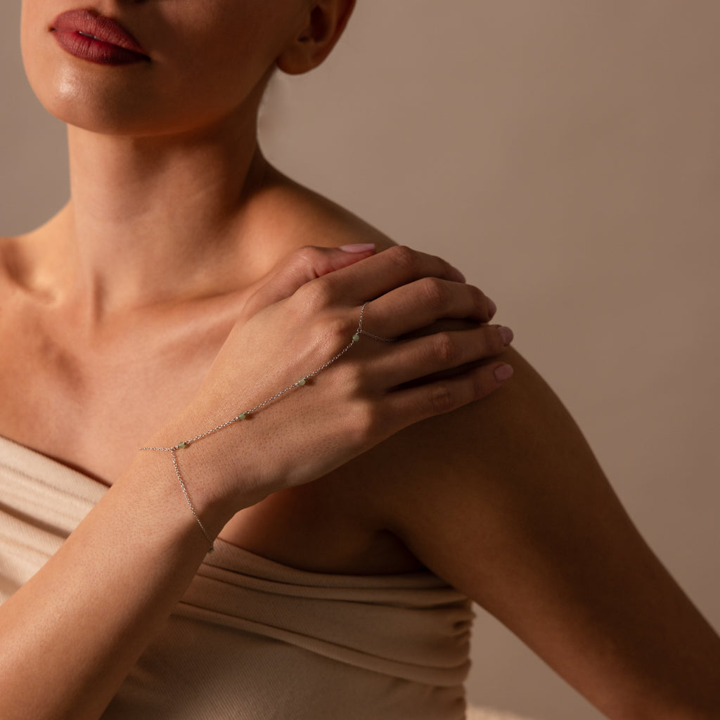 Woman in a beige off-shoulder top elegantly showcases the Jade Hand Chain Bracelet in Silver, featuring delicate shimmering jade beads on her hand and wrist.