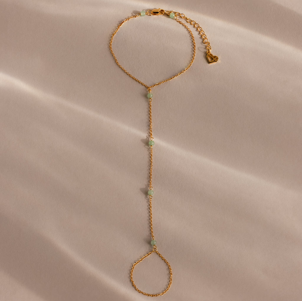 The Jade Hand Chain Bracelet in 18K Gold features jade beads and a heart charm, shown on a soft beige surface.