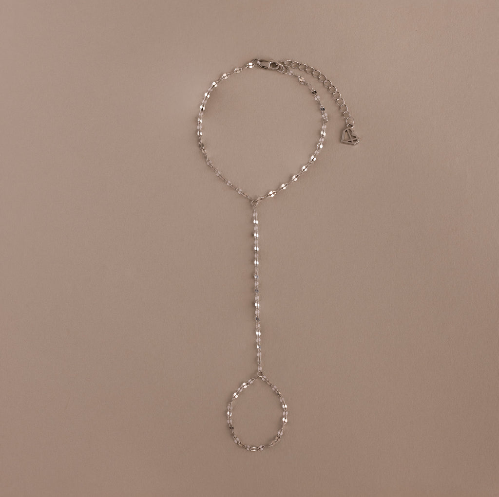 The Sparkle Hand Chain Bracelet in Silver features mirror cut links, a linked finger loop, and an adjustable clasp, beautifully displayed on a beige background.