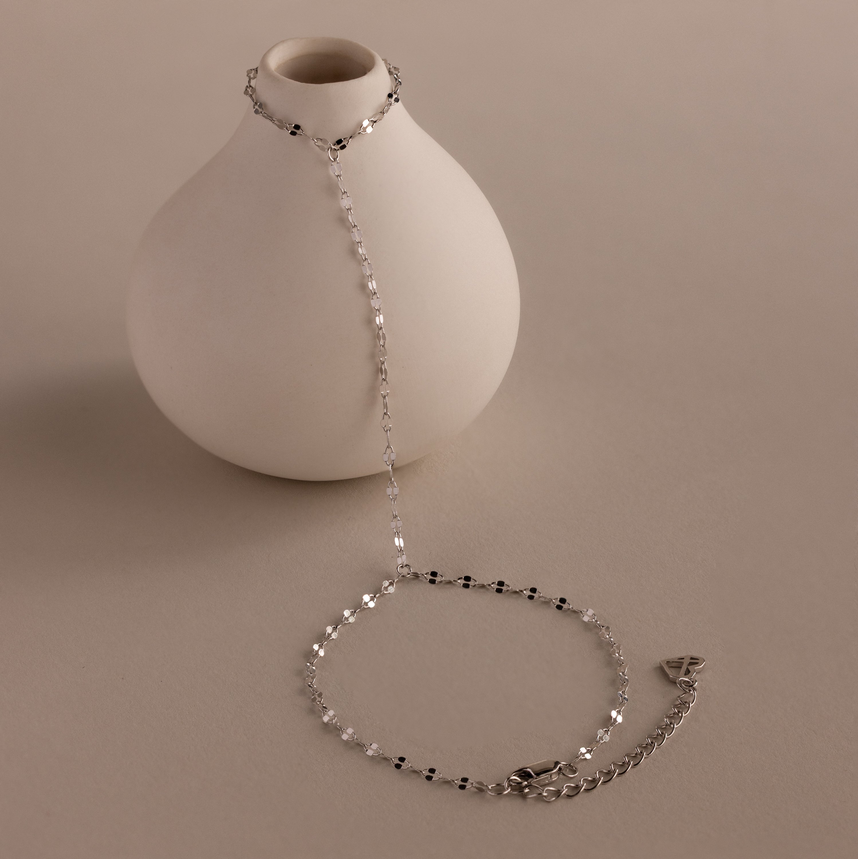 The Sparkle Hand Chain Bracelet in Silver, featuring mirror-cut links and black and clear beads, is draped over a round white vase on a neutral surface.