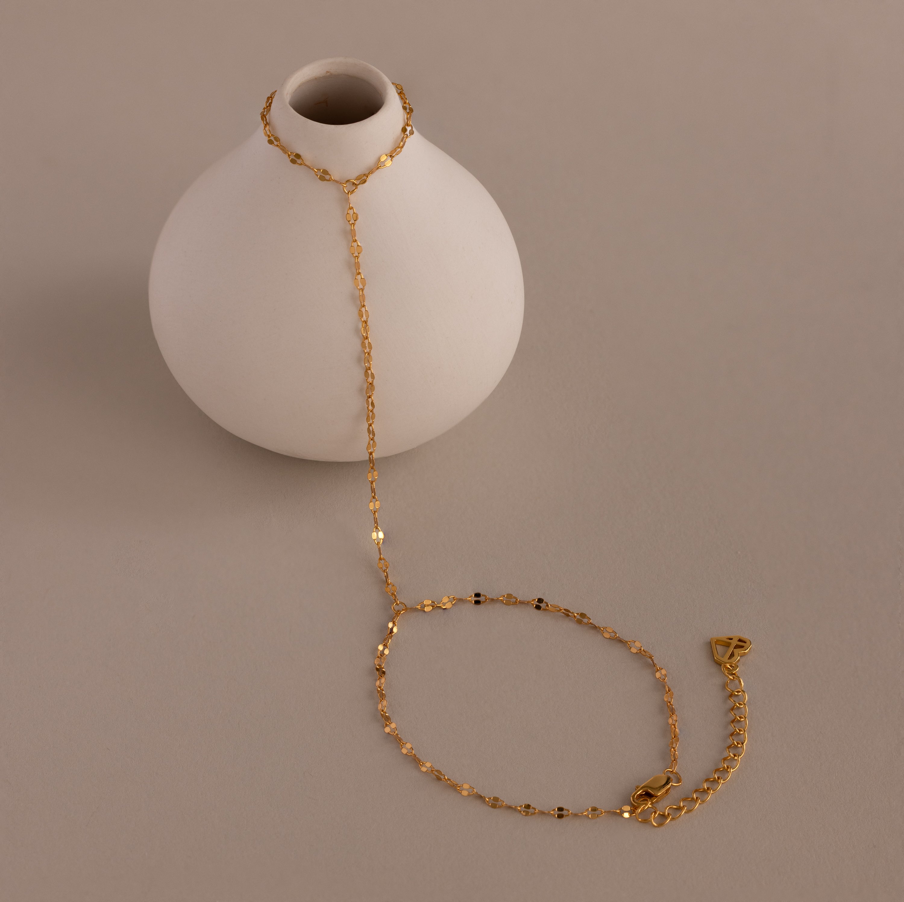 The Sparkle Hand Chain Bracelet, featuring delicate mirror-cut gold links, is elegantly draped over a small round white vase on a neutral background.