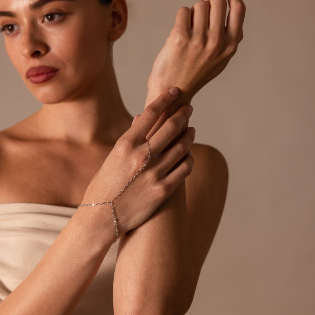 A woman touches her wrist while wearing the Sparkle Hand Chain Bracelet in Silver, featuring delicate mirror-cut links, set against a neutral background.
