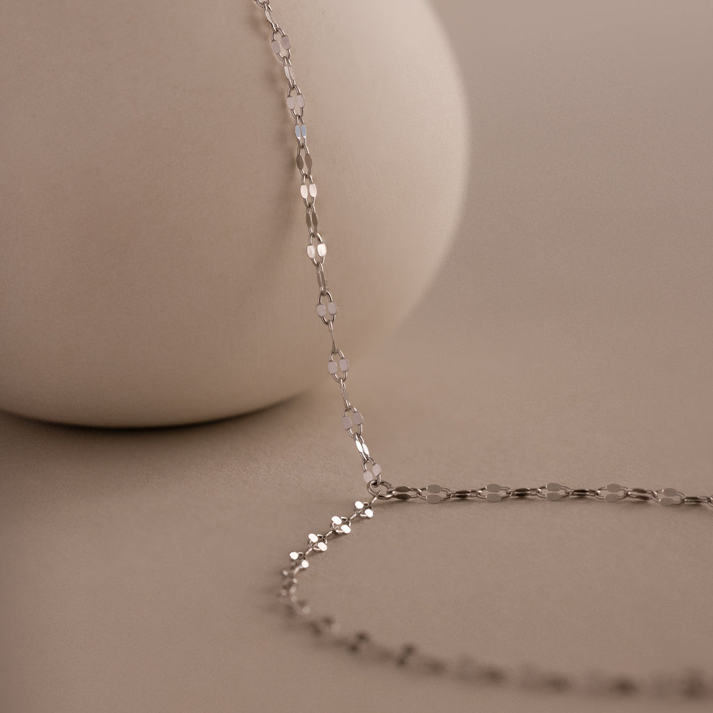 The Sparkle Hand Chain Bracelet in Silver, featuring mirror-cut links, is elegantly draped over a smooth, round beige surface on a soft beige background.