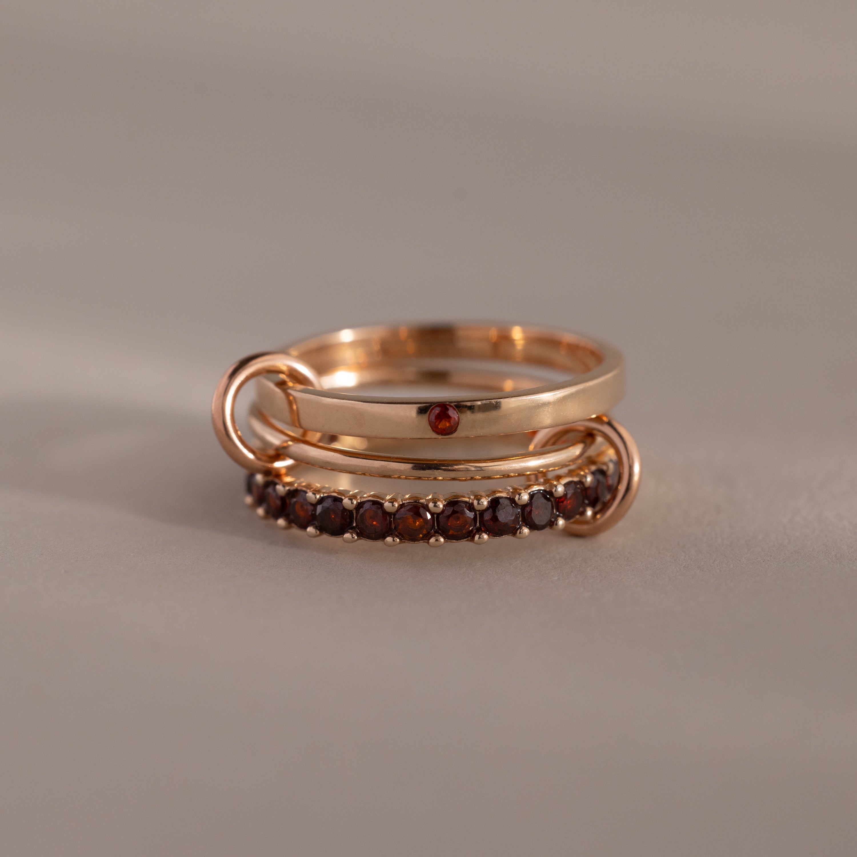 The Triple Linked Garnet Ring Set features two stacked gold rings: one with a single small red stone and another eternity band adorned with a row of small red garnet gemstones.