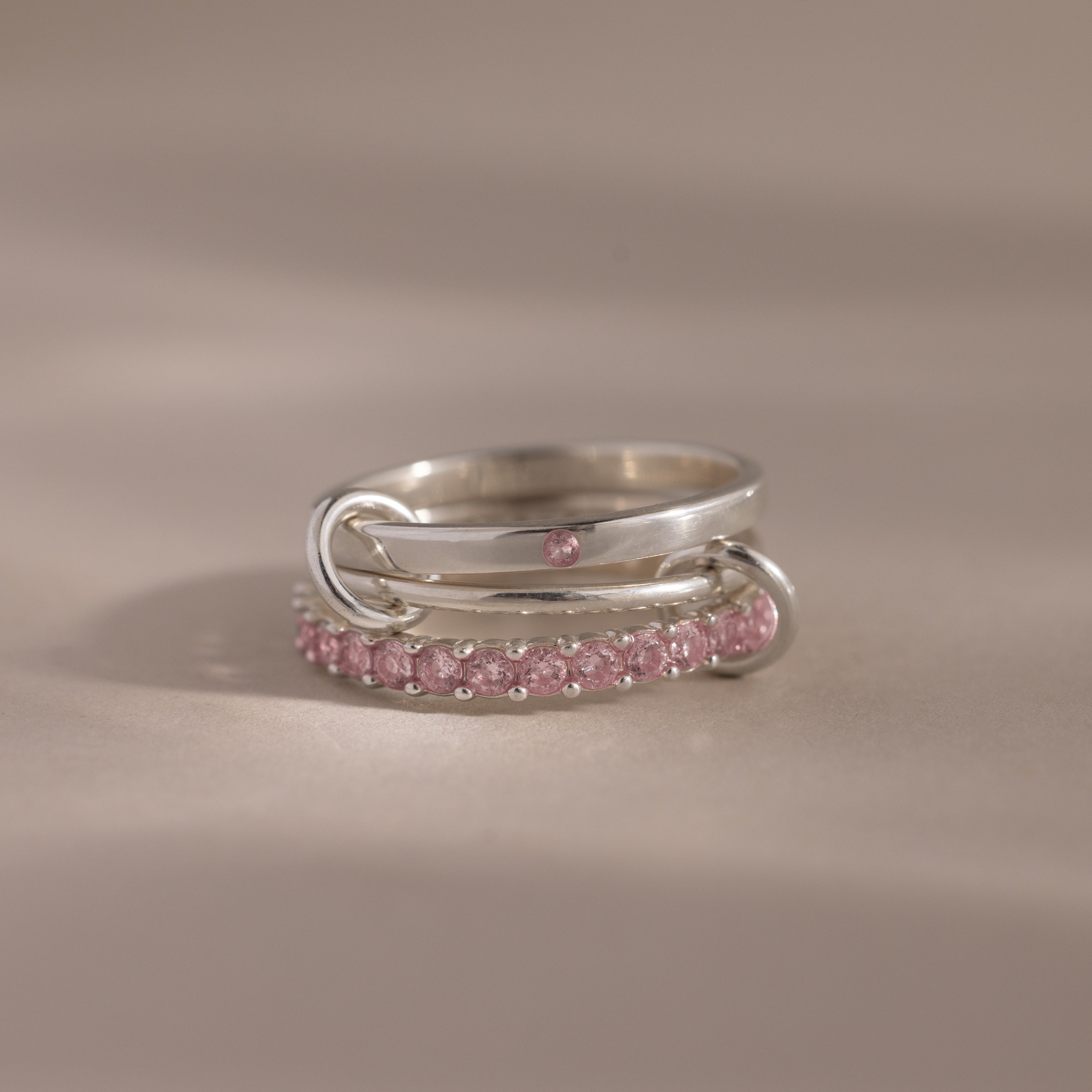 The Triple Linked Pink Tourmaline Ring Set features intertwined silver bands and a single pink tourmaline stone on top, beautifully displayed on a beige surface.