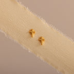 Dainty gold cross stud earrings displayed on a neutral ribbon fabric.