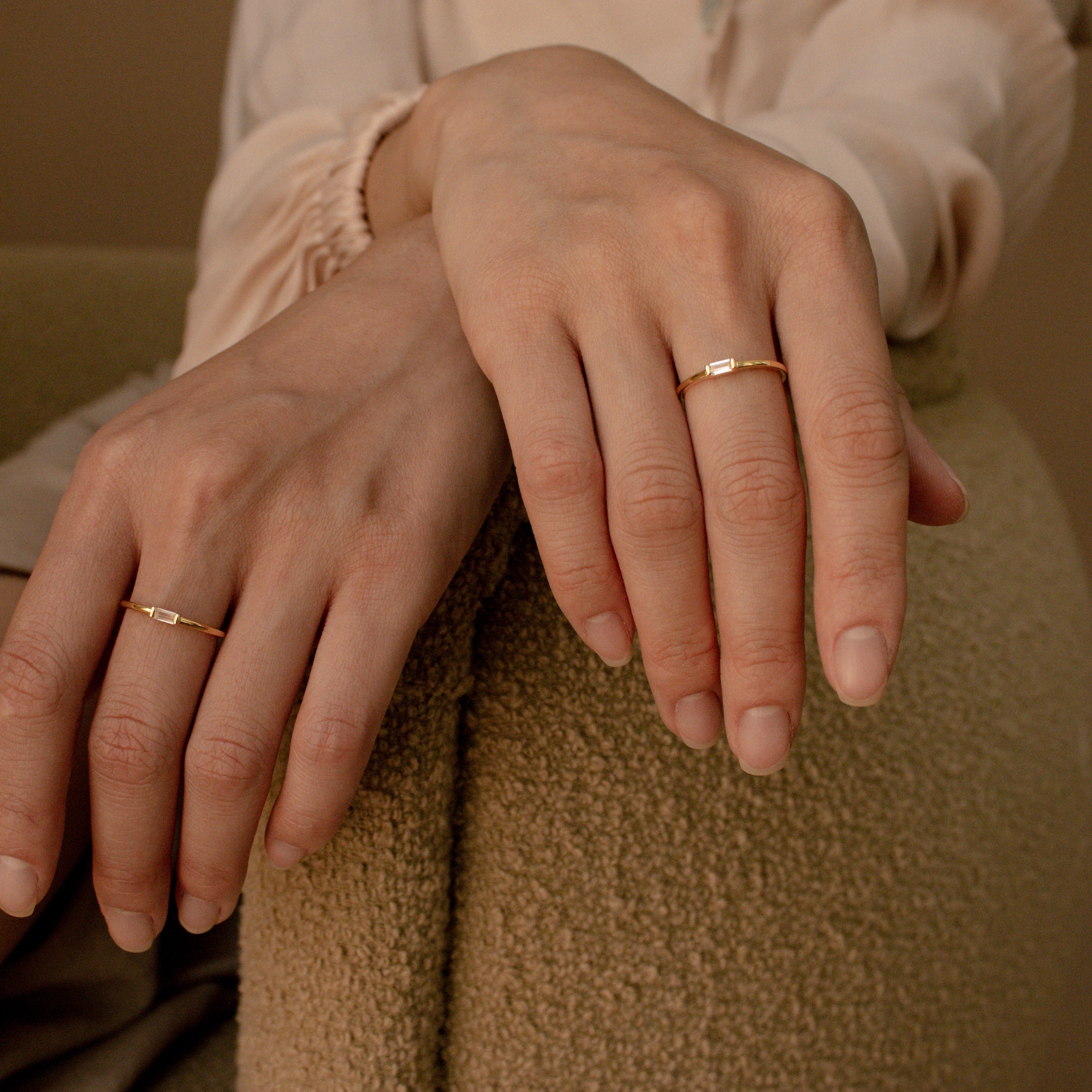 Two hands adorned with delicate gold rings, including the Baguette Diamond Ring in Silver set east-west, rest on an armchair while wearing a light long-sleeved blouse.