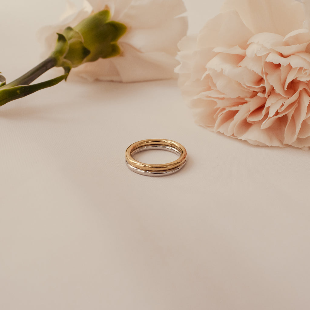 The Minimal Stacking Ring in 18K Gold is shown on a white surface with light pink carnations, ideal for creating a chic, minimal finger stack look.