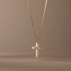 The Baroque Pearl Cross Necklace in 18K Gold features a white freshwater pearl cross pendant on a gold chain, elegantly displayed against a neutral beige background.