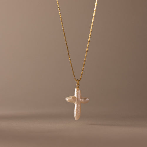 The Baroque Pearl Cross Necklace in 18K Gold features a white freshwater pearl cross pendant on a gold chain, elegantly displayed against a neutral beige background.