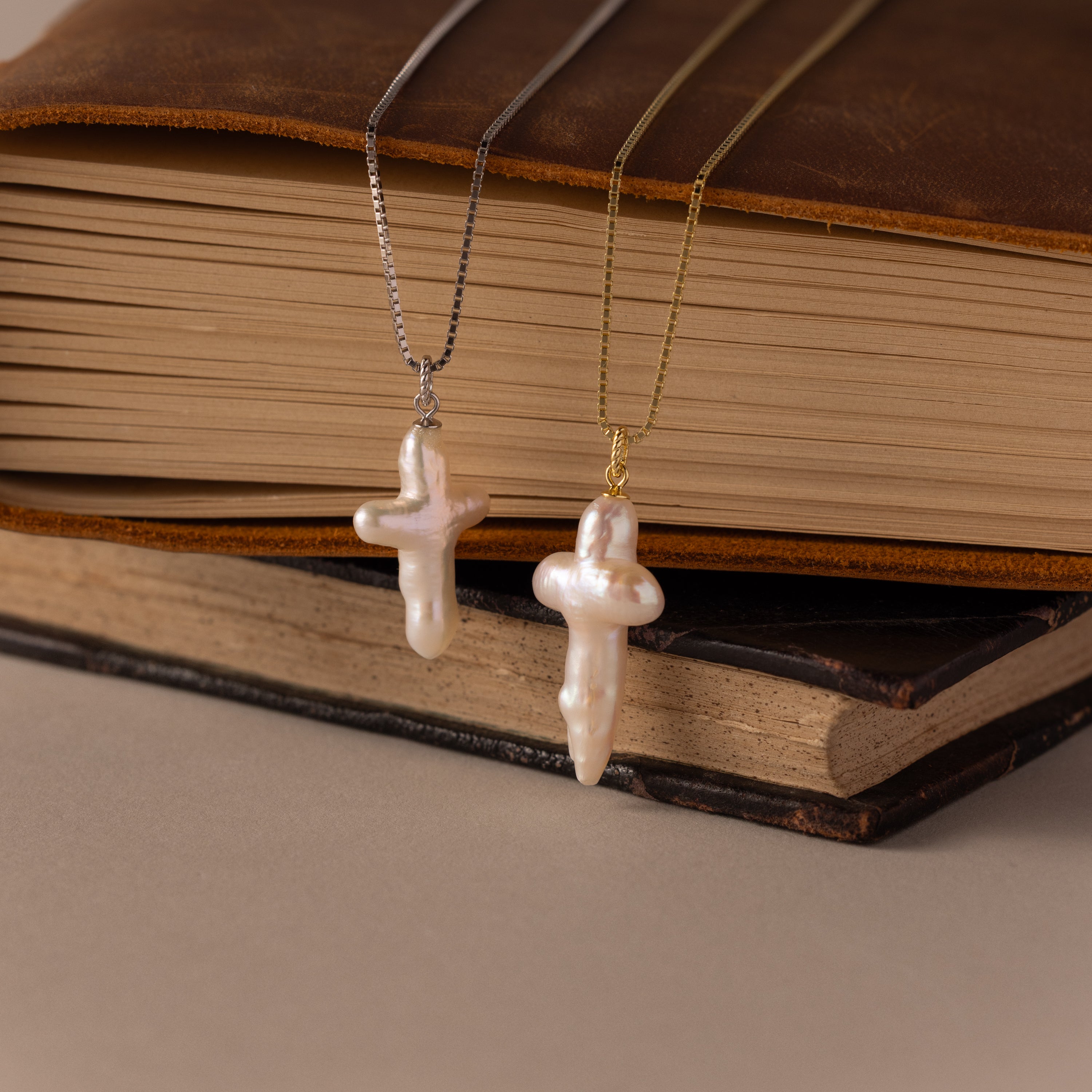 Two Baroque Pearl Cross Necklaces in Silver with minimalist chains rest gracefully against the pages of two stacked books.