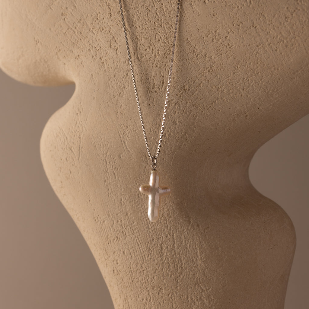 The Baroque Pearl Cross Necklace in Silver, featuring a minimalist chain and freshwater pearl cross pendant, is displayed on a textured beige mannequin bust, adding elegant style to any outfit.