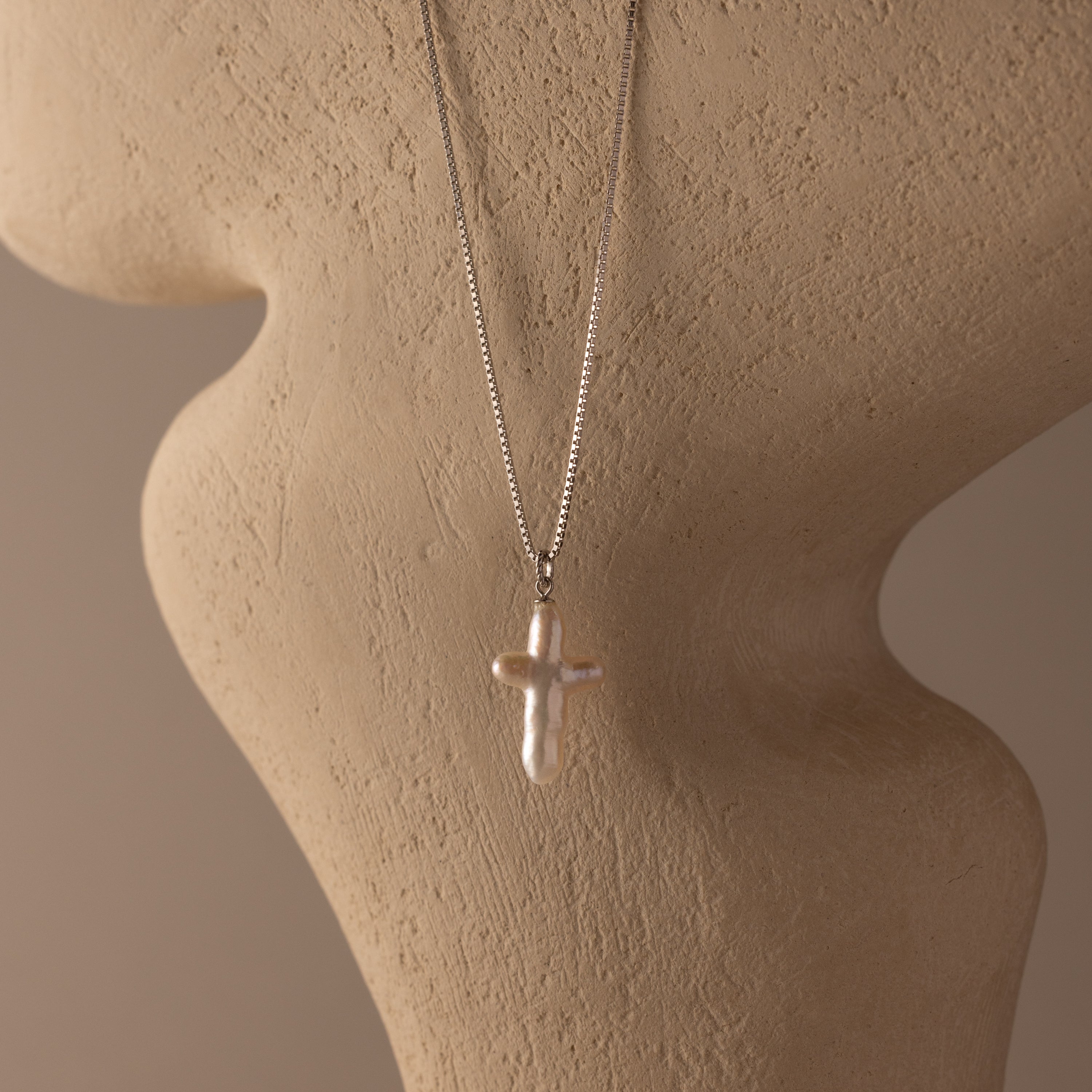 The Baroque Pearl Cross Necklace in Silver, featuring a minimalist chain and freshwater pearl cross pendant, is displayed on a textured beige mannequin bust, adding elegant style to any outfit.