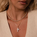 A woman wears a freshwater pearl necklace and the Baroque Pearl Cross Necklace in 18K Gold, styled with a minimalist gold chain and a beige knit top.