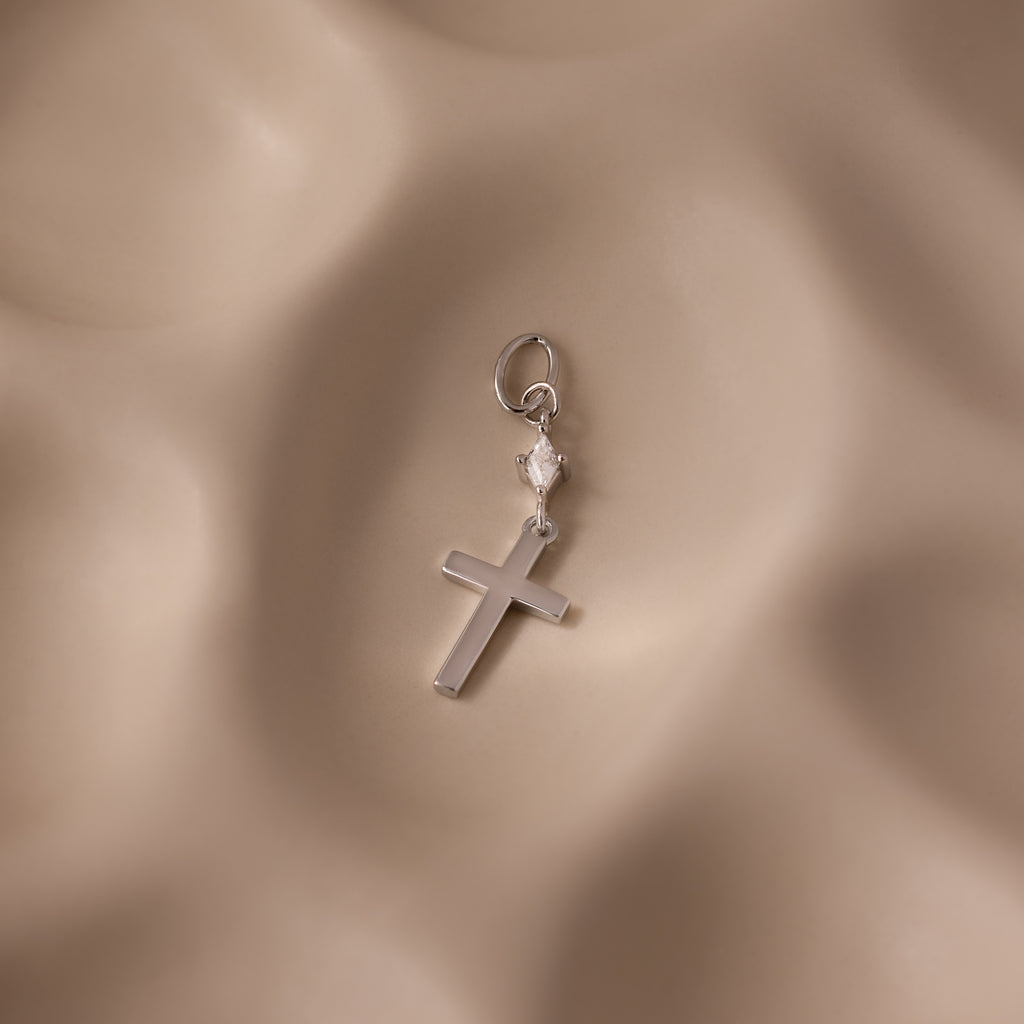 The Diamond Drop Cross Charm in Silver features a delicate cross pendant with a sparkling diamond drop and a small heart, displayed on a soft beige textured background.