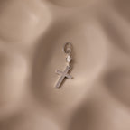 The Diamond Drop Cross Charm in Silver features a delicate cross pendant with a sparkling diamond drop and a small heart, displayed on a soft beige textured background.
