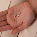 A hand holds the Diamond Drop Cross Charm in Silver against a beige background, its delicate design exuding subtle elegance reminiscent of a sparkling diamond.