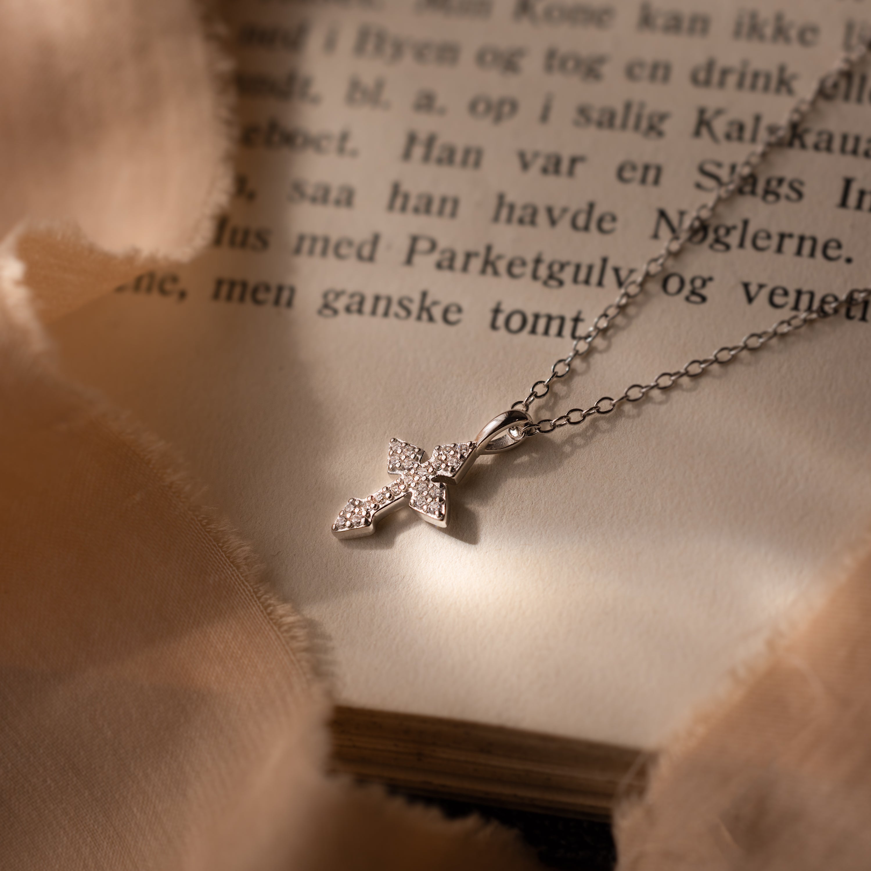 The Vintage Cross Necklace in Silver, featuring a pavé finish and delicate diamonds, rests on the open pages of a book, surrounded by soft fabric.