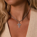 A woman wears the Majesty Cross Necklace in Silver adorned with sparkling stones over a beige knit top.