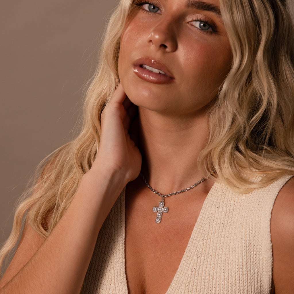 Woman with long blonde hair wears a cream sleeveless top and the Majesty Cross Necklace in Silver, sparkling as she touches her neck.