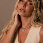 Woman with long blonde hair wears a cream sleeveless top and the Majesty Cross Necklace in Silver, sparkling as she touches her neck.
