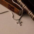 The Majesty Cross Necklace in Silver, adorned with sparkling stones, is displayed next to two vintage books on a beige surface.