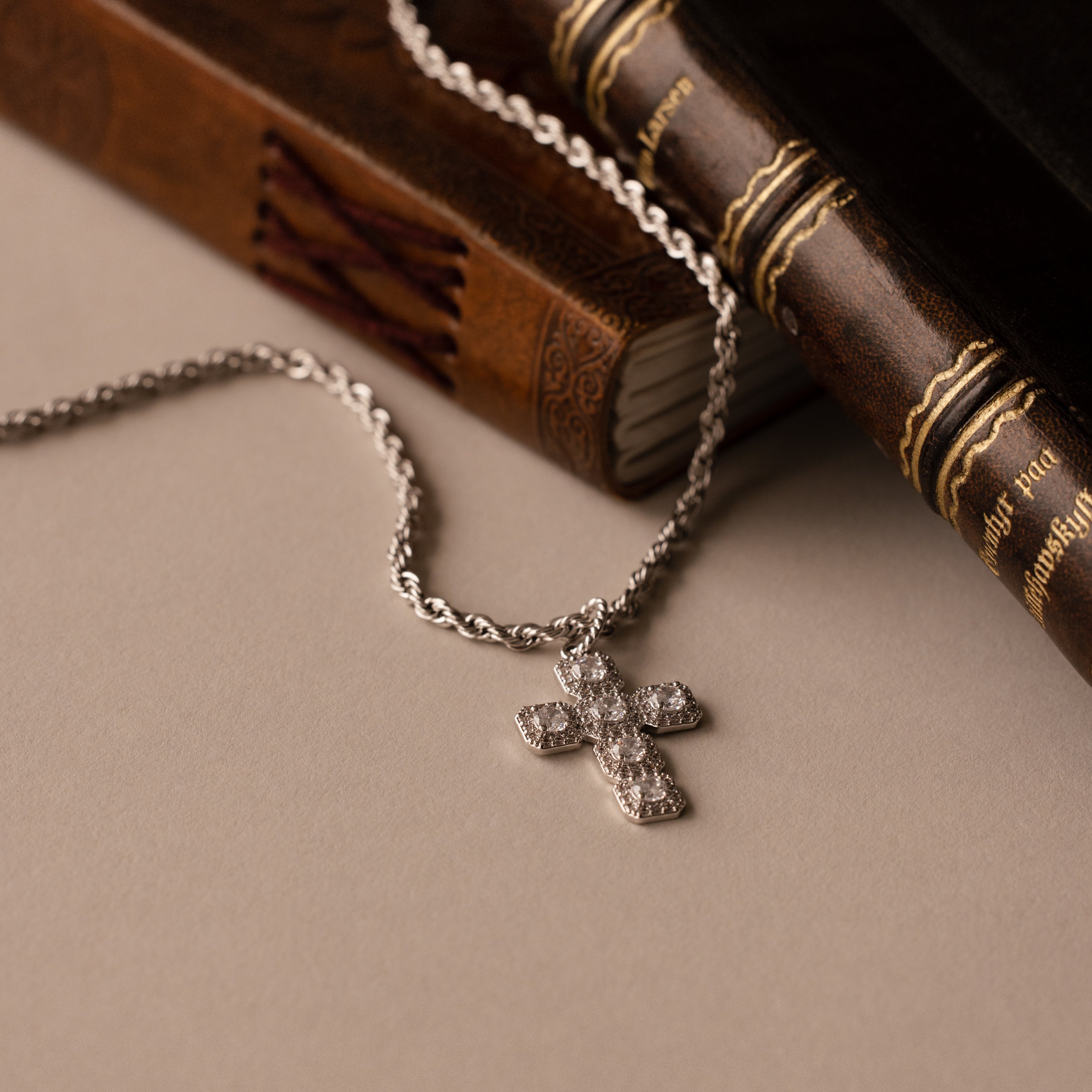 The Majesty Cross Necklace in Silver, adorned with sparkling stones, is displayed next to two vintage books on a beige surface.