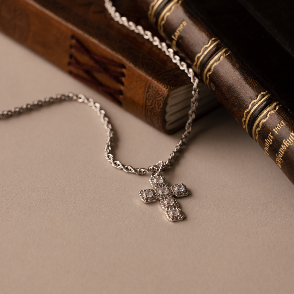 The Majesty Cross Necklace, adorned with sparkling diamonds, rests on vintage leather-bound books atop a beige surface, beautifully highlighting the enduring elegance of faith-inspired jewelry.