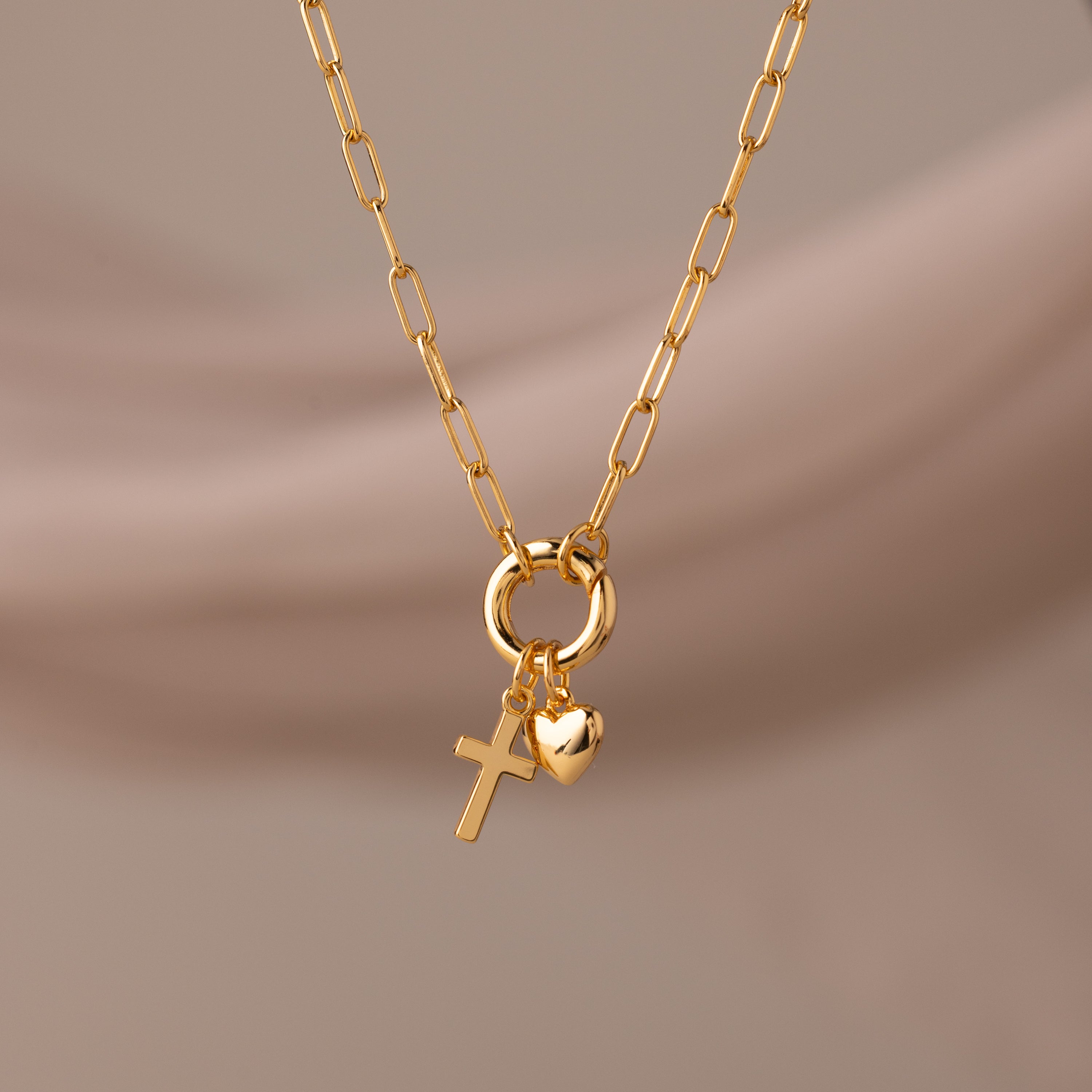 Cross & Heart Charm Bracelet in 18K Gold, featuring cross and heart charms on a stylish paperclip chain, displayed against a soft beige background.