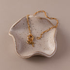 The Cross & Heart Charm Bracelet, featuring cross and heart charms, is displayed in a speckled ceramic dish on a beige background.