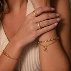 A woman’s hands showcase a Cross & Heart Charm Bracelet in 18K Gold and gold rings, styled against a beige sleeveless top.