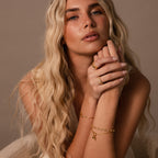Woman with long blonde hair wearing gold jewelry, including the Cross & Heart Charm Bracelet, poses with her hands near her face against a neutral background.