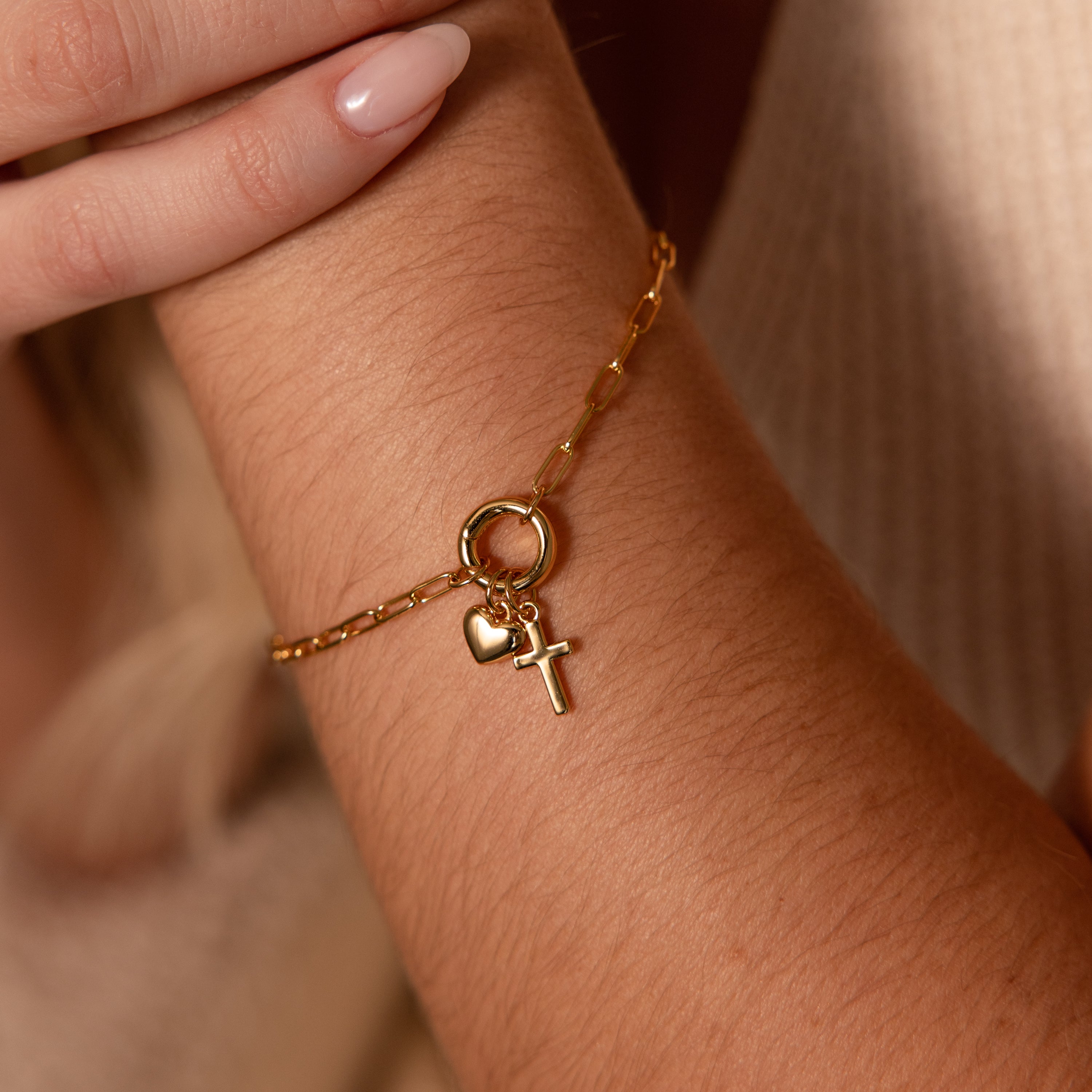 The Cross & Heart Charm Bracelet in 18K Gold features a minimalist paperclip chain with cross and heart charms, elegantly wrapped around the wrist.